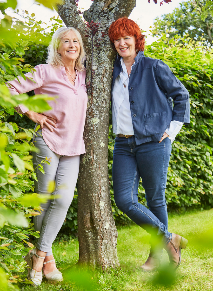 Susi Lennox and Sarah Brooks. Founders of the YES YES Company standing in an English garden leaning against a tree looking happy and relaxed